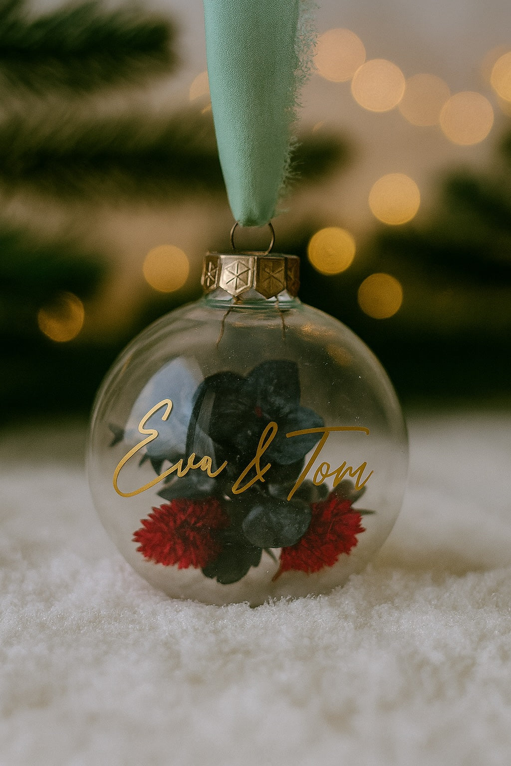 Personalisierte Weihnachtskugel aus Glas mit goldenem Kranz und grünen Chiffon-Aufhänger, gefüllt mit roten Phalaris-Trockenblumen und grünem Eukalyptus, Variante ‚Winter Flame‘. Mit Personalisierung Eva&Tom in goldener Schrift.