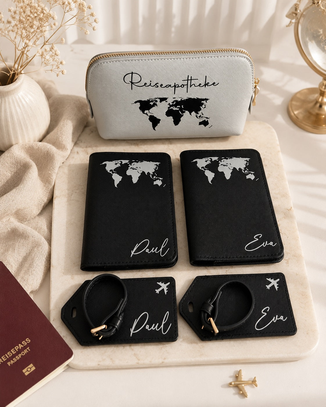 Personalisiertes Reiseset mit schwarzer Reisepasshülle und Kofferanhängern mit weißem Weltkarten-Aufdruck sowie hellgrauer Reiseapotheke mit schwarzem Aufdruck, dekorativ arrangiert mit Reisepass auf hellem Hintergrund.