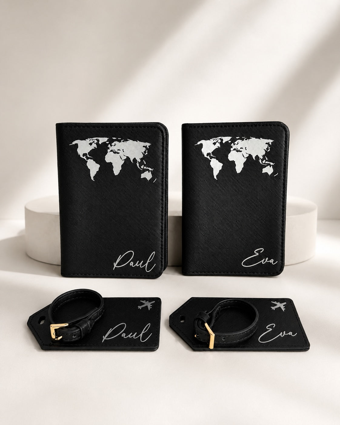 Personalisiertes Reisepasshüllen-Set in Schwarz mit weißem Weltkarten-Aufdruck, bestehend aus zwei Reisepasshüllen und passenden Kofferanhängern mit den Namen Paul und Eva auf hellem, neutralem Hintergrund.