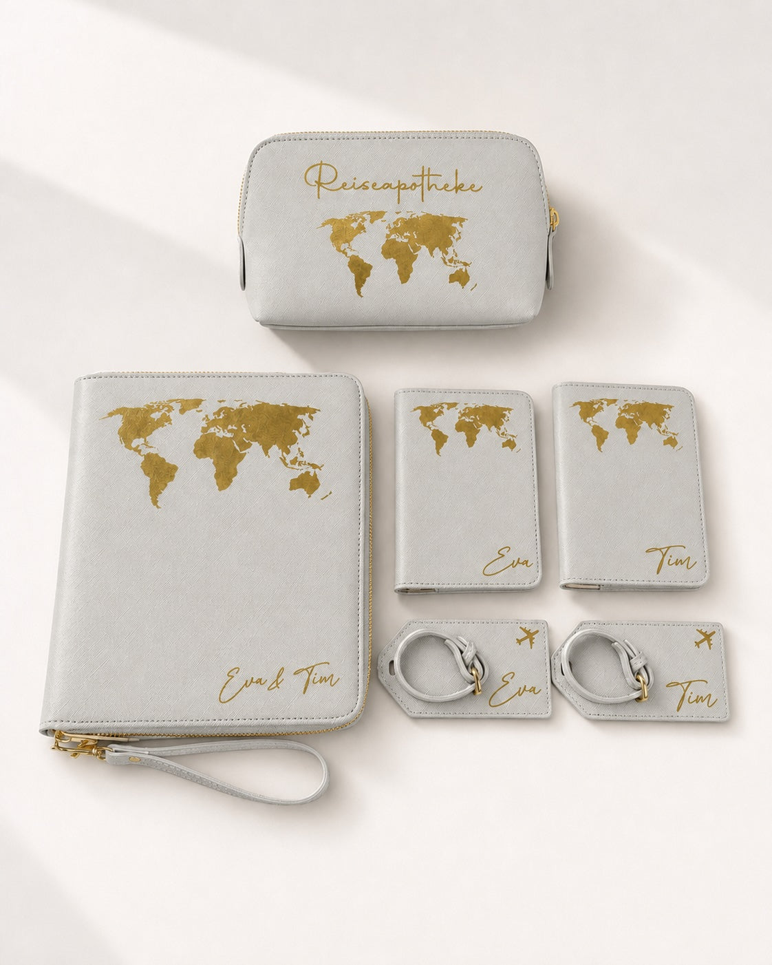 Personalisiertes Reiseset in Hellgrau mit goldenen Weltkarten- und Schriftaufdrucken, bestehend aus Reiseapotheke, Reiseorganizer, Reisepasshüllen und Kofferanhängern mit den Namen Eva und Tim auf hellem, neutralem Hintergrund.