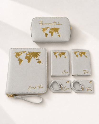 Personalisiertes Reiseset in Hellgrau mit goldenen Weltkarten- und Schriftaufdrucken, bestehend aus Reiseapotheke, Reiseorganizer, Reisepasshüllen und Kofferanhängern mit den Namen Eva und Tim auf hellem, neutralem Hintergrund.
