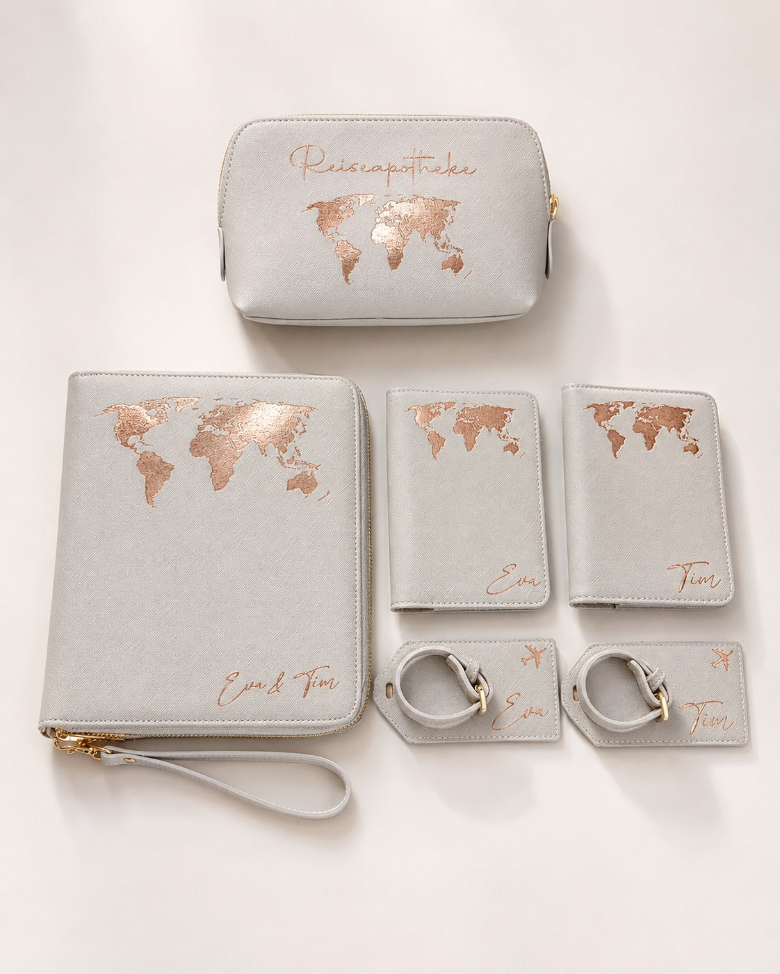 Personalisiertes Reiseset in Hellgrau mit roségoldenem, leicht schimmerndem Weltkarten-Aufdruck, bestehend aus Reiseorganizer, Reiseapotheke, Reisepasshüllen und Kofferanhängern mit individuellen Namen auf hellem, neutralem Hintergrund.