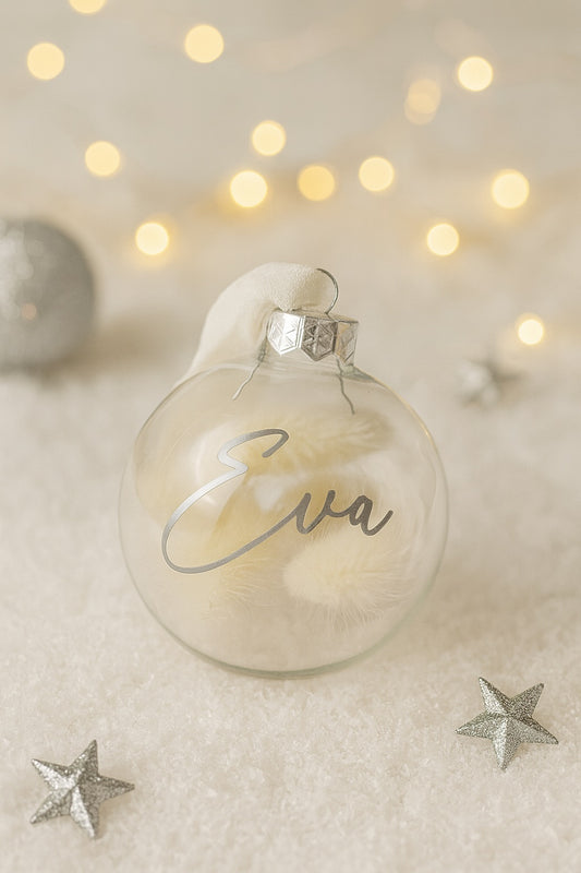 Eine transparente Weihnachtskugel mit weißem Stoffband liegt auf Kunstschnee. Im Inneren befinden sich cremefarbene Trockenblumen. Auf der Kugel steht in silberner Schrift der Name „Eva“. Um die Kugel herum liegen silberne Sterne, im Hintergrund leuchten unscharfe warme Lichter.