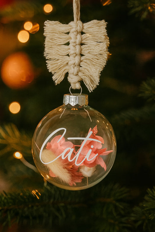 Personalisierte gläserne Weihnachtskugel mit der Aufschrift Cati, gefüllt mit rosa Blüten und einem cremefarbenen Trockenblumen-Element. Die Kugel hängt an einem Makramee-Band vor einem geschmückten Weihnachtsbaum mit goldenen Lichtern im Hintergrund.