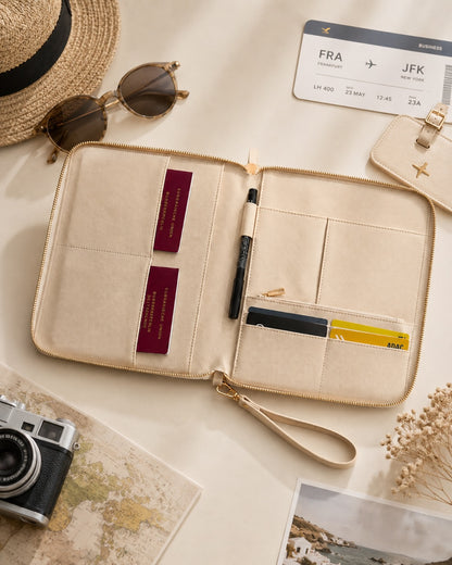 Geöffneter beiger Reise-Organizer mit Kartenfächern und Stift, daneben Reisepass, Reiseunterlagen und Accessoires wie Kamera, Sonnenbrille und Hut auf einem hellen, neutralen Reise-Flatlay.