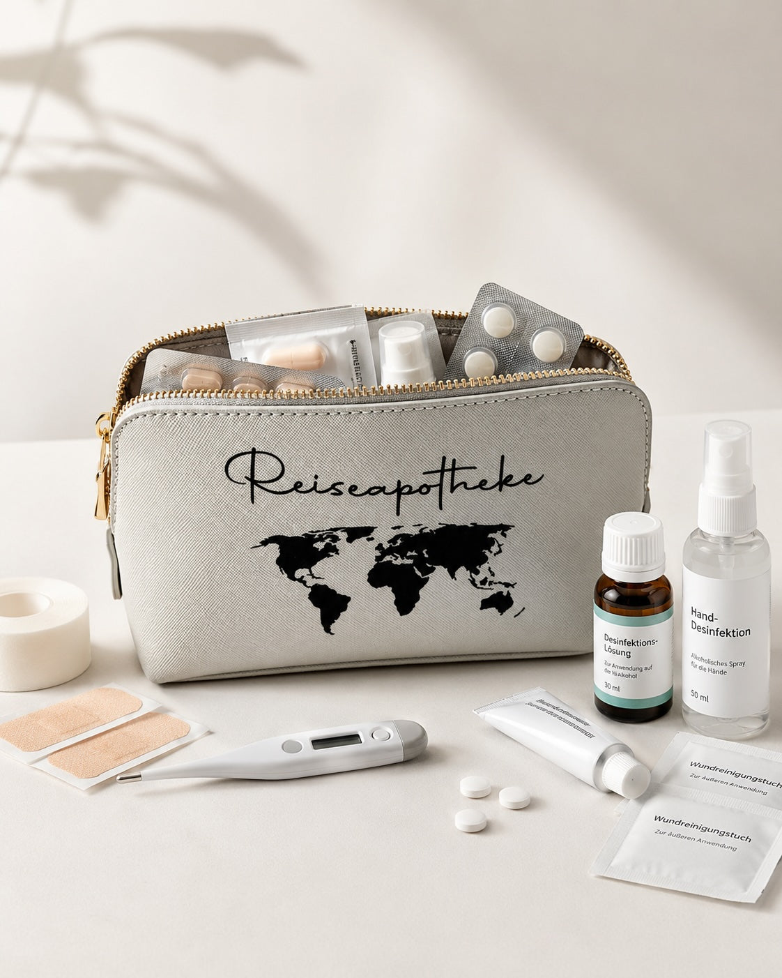 Modernes Produktbild einer hellgrauen Reiseapotheke-Tasche mit schwarzem Aufdruck, geöffnet und mit Medikamenten, Pflastern, Thermometer und Desinfektion vor hellem, neutralem Hintergrund.