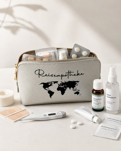 Modernes Produktbild einer hellgrauen Reiseapotheke-Tasche mit schwarzem Aufdruck, geöffnet und mit Medikamenten, Pflastern, Thermometer und Desinfektion vor hellem, neutralem Hintergrund.