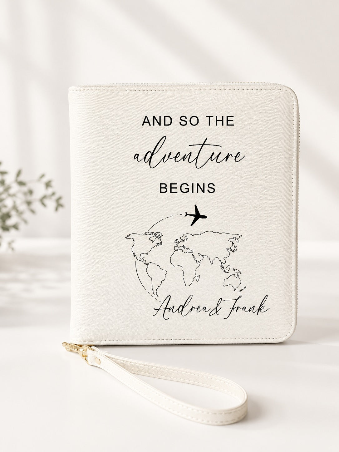 Beiger personalisierter Reiseorganizer mit schwarzem Schriftzug „And so the adventure begins“, Weltkarten-Motiv, Flugzeug und den Namen Andrea und Frank vor hellem, neutralem Hintergrund.