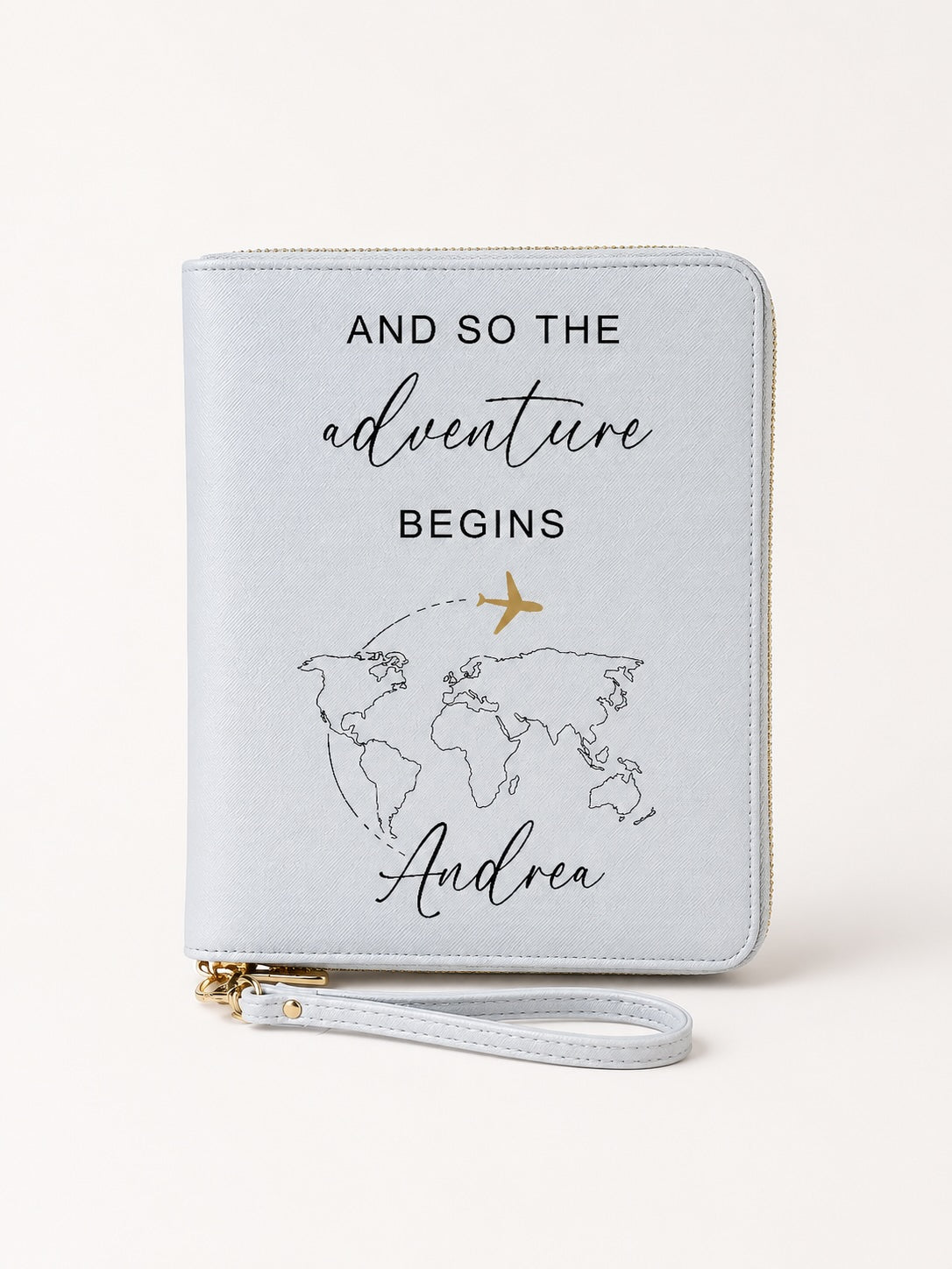 Grauer personalisierter Reiseorganizer mit schwarzem Schriftzug „And so the adventure begins“, Weltkarten-Motiv, goldenem Flugzeug und dem Namen Andrea vor hellem, einfarbigem Hintergrund.