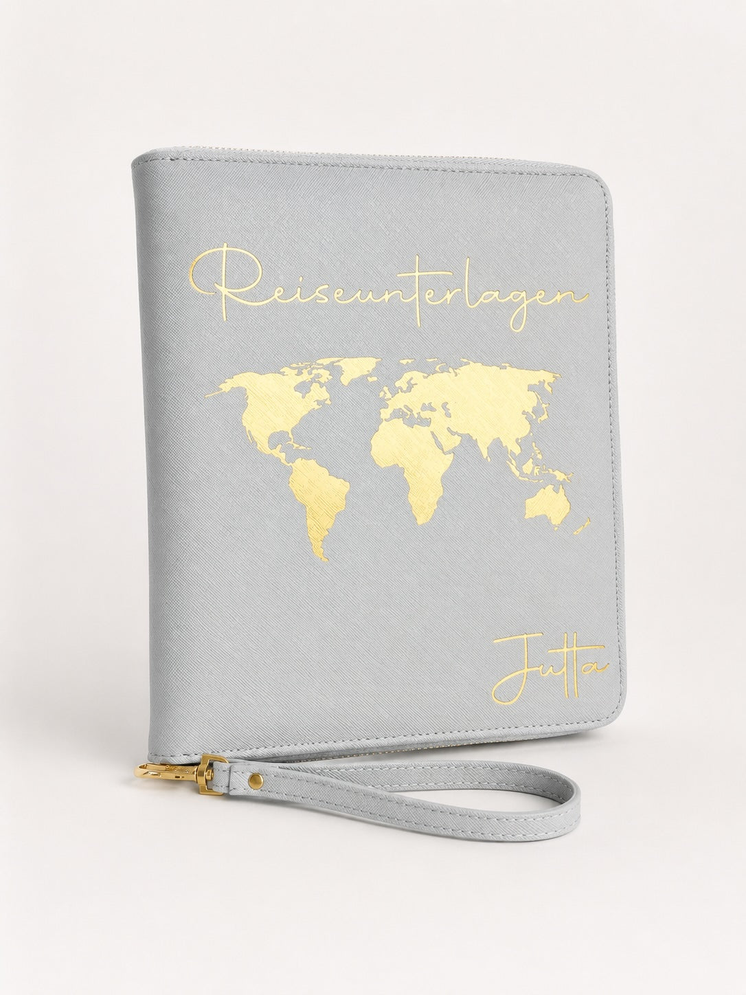 Grauer Reiseorganizer mit vollständig goldenem Aufdruck, Weltkarte, dem Schriftzug „Reiseunterlagen“ und dem Namen Jutta vor hellem, neutralem Hintergrund.