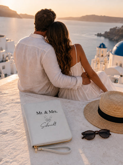 Personalisierter hellgrauer Reiseorganizer „Mr. & Mrs. Schmidt“ mit schwarzem Handmotiv und goldenen Details vor romantischer Flitterwochenkulisse.
