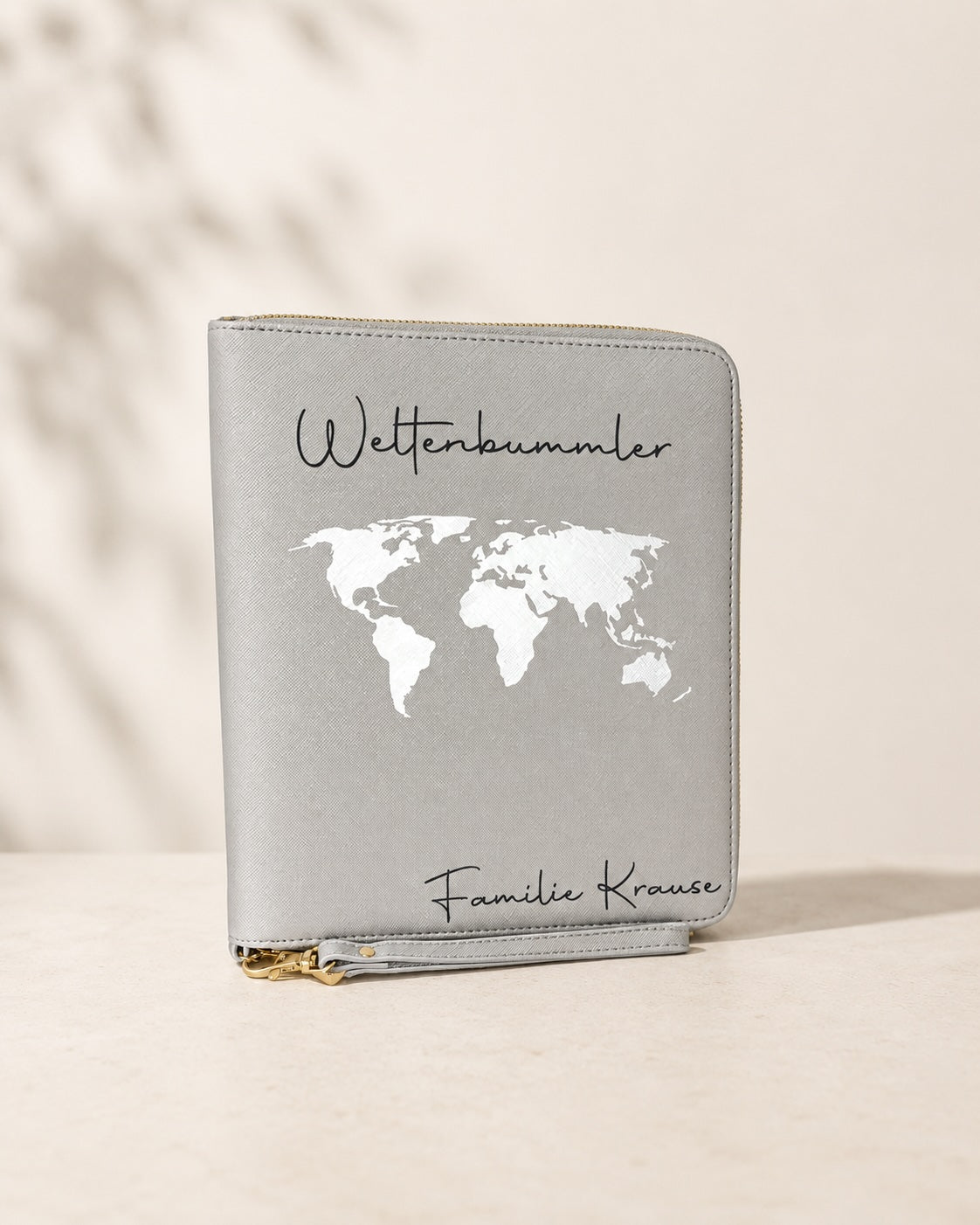 Grauer personalisierter Reiseorganizer mit weißer Weltkarte, schwarzem Schriftzug „Weltenbummler“ und der Personalisierung „Familie Krause“ vor hellem neutralem Hintergrund.
