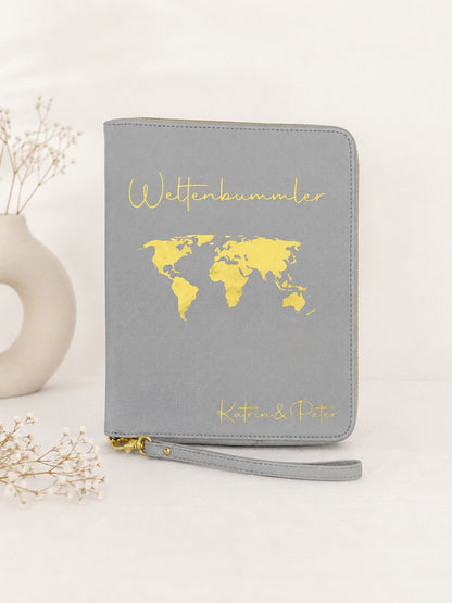 Grauer personalisierter Reiseorganizer mit goldenem Schriftzug „Weltenbummler“, Weltkarte und den Namen Katrin und Peter vor hellem neutralem Hintergrund.