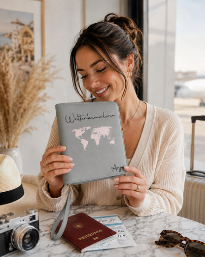 Lächelnde Frau hält einen grauen personalisierten Reiseorganizer mit rosafarbener Weltkarte, dem Schriftzug „Weltenbummlerin“ und dem Namen Anja. Im Hintergrund liegen Reisepass, Flugtickets, Kamera, Sonnenbrille und Koffer.