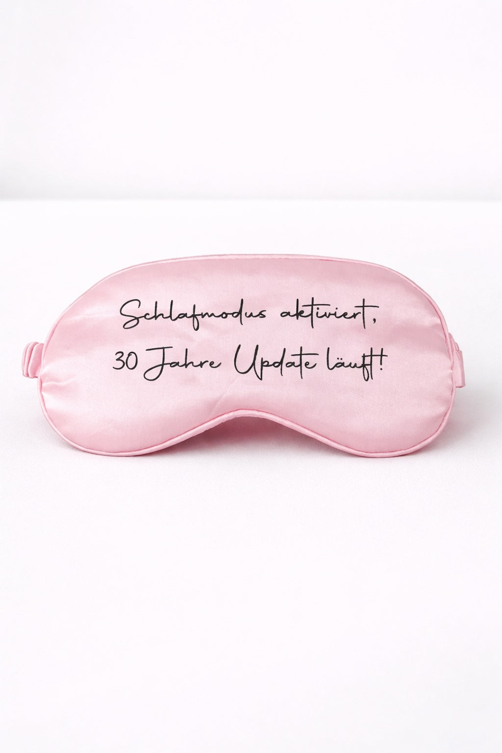 Rosa Schlafmaske mit Aufdruck ‚Schlafmodus aktiviert, 30 Jahre Update läuft!‘ vor hellem, minimalistischem Hintergrund