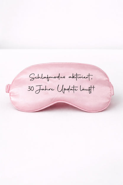 Rosa Schlafmaske mit Aufdruck ‚Schlafmodus aktiviert, 30 Jahre Update läuft!‘ vor hellem, minimalistischem Hintergrund
