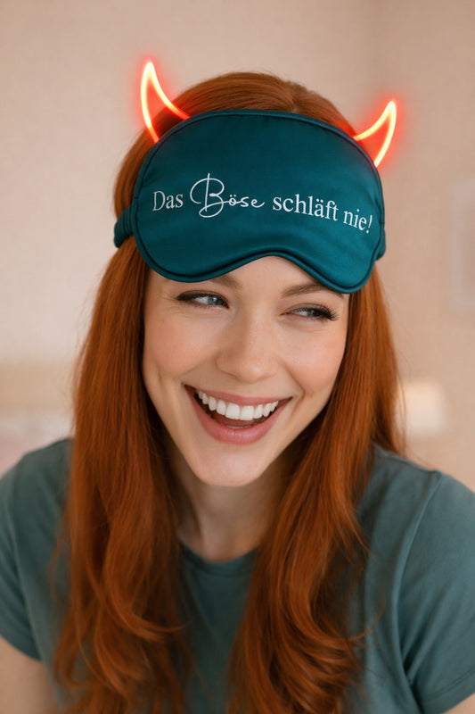 Junge Frau mit verschmitztem Lächeln trägt eine dunkelgrüne Satin-Schlafmaske mit Aufdruck ‚Das Böse schläft nie‘ und leuchtenden Teufelshörnern