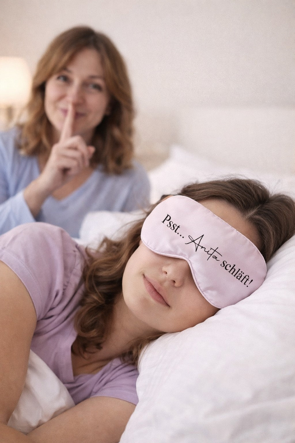 Junge Frau schläft mit personalisierter Schlafmaske „Psst… Anita schläft“, im Hintergrund lächelnde Mutter mit Leise-Geste