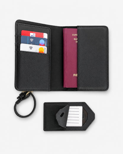 Offenes schwarzes Reiseportemonnaie auf hellem, neutralem Hintergrund. Im Inneren befinden sich Kartenfächer mit Kreditkarten sowie ein bordeauxfarbener Reisepass in der rechten Einsteckhülle. Links ist eine Handschlaufe befestigt. Darunter liegt ein schwarzer Gepäckanhänger mit Einsteckkarte, der teilweise geöffnet ist. Produktfoto im minimalistischen Stil für einen Online-Shop.