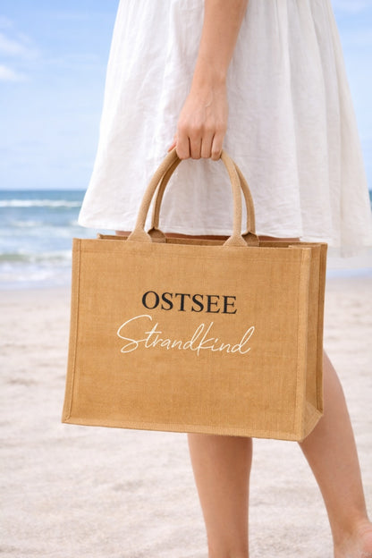 Frau trägt eine große Jute Strandtasche mit Aufdruck „OSTSEE Strandkind“ am Strand vor dem Meer.