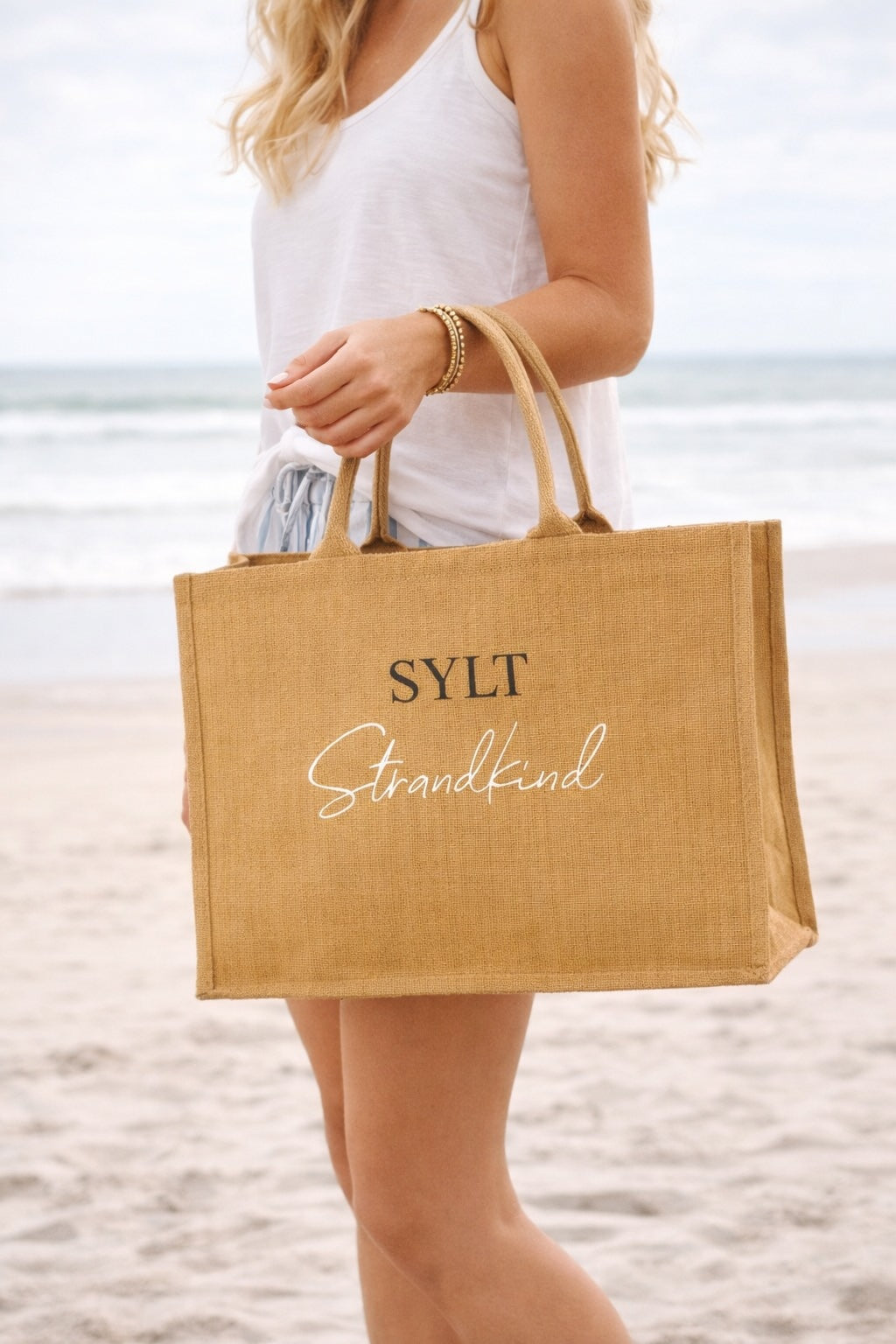 Frau trägt eine große Jute-Strandtasche mit dem Aufdruck „SYLT Strandkind“ am Strand vor dem Meer.