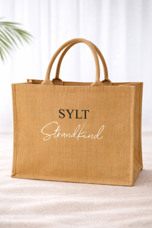 Jute Strandtasche in Naturfarbe mit Aufdruck „SYLT Strandkind“ in schwarzer und weißer Schrift vor hellem, neutralem Hintergrund.
