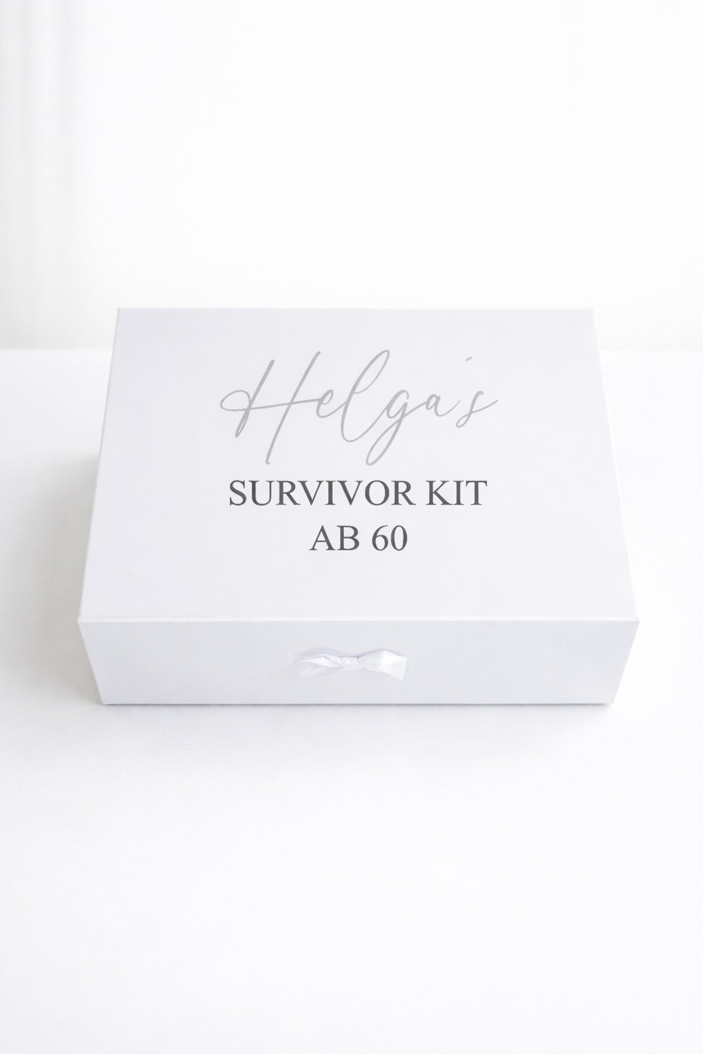 Weiße personalisierte Geschenkbox ‚Helga’s Survivor Kit ab 60‘ mit Schleife auf hellem, minimalistischem Hintergrund