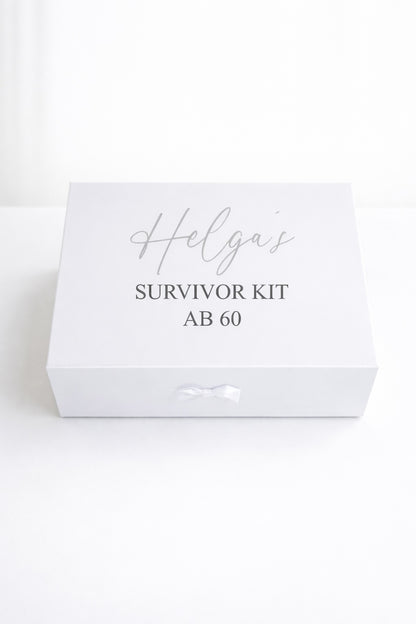 Weiße personalisierte Geschenkbox ‚Helga’s Survivor Kit ab 60‘ mit Schleife auf hellem, minimalistischem Hintergrund