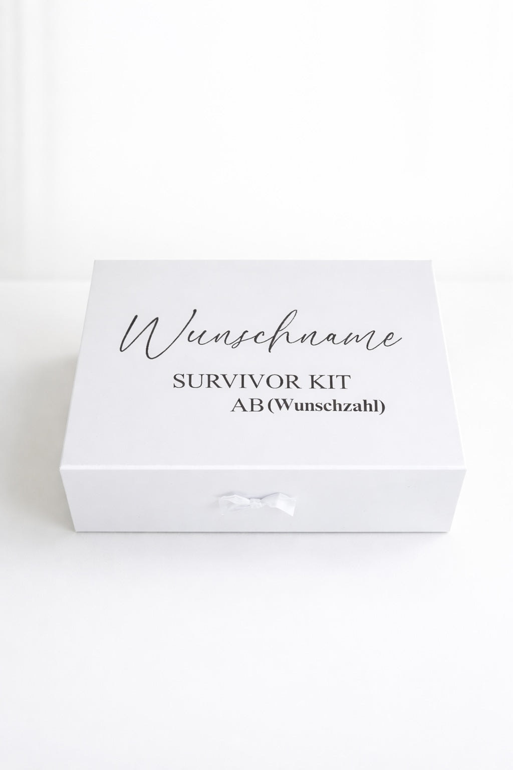 Weiße personalisierte Geschenkbox ‚Survivor Kit ab (Wunschzahl)‘ mit Schleife auf hellem, minimalistischem Hintergrund