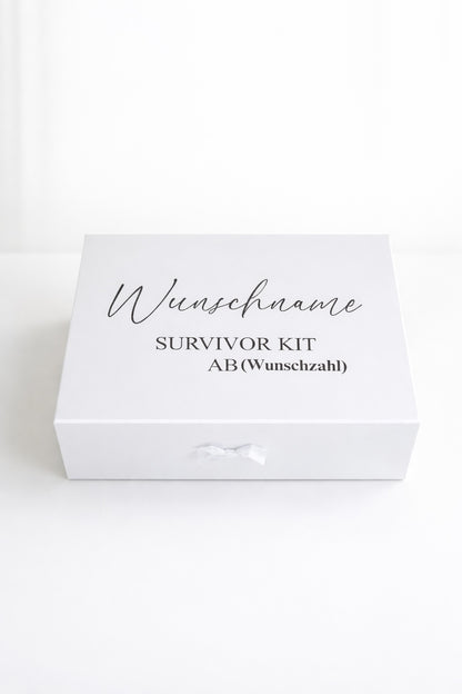 Weiße personalisierte Geschenkbox ‚Survivor Kit ab (Wunschzahl)‘ mit Schleife auf hellem, minimalistischem Hintergrund