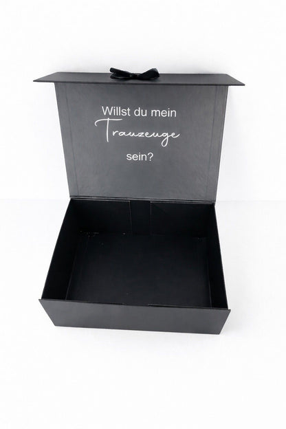 Offene schwarze Geschenkbox mit der Aufschrift ‚Willst du mein Trauzeuge sein?‘ auf hellem, minimalistischem Hintergrund.