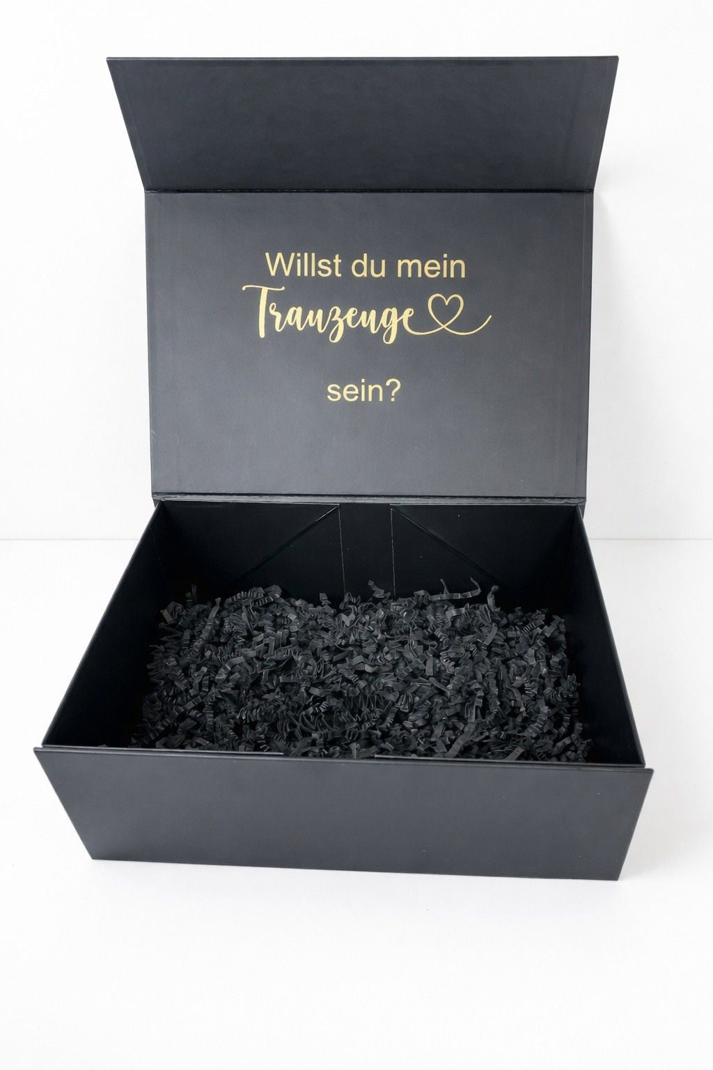 Offene schwarze Geschenkbox mit Füllpapier und goldener Aufschrift ‚Willst du mein Trauzeuge sein?‘ auf hellem, minimalistischem Hintergrund