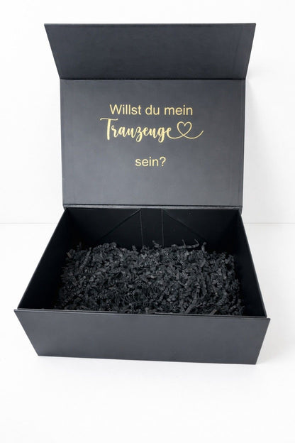 Offene schwarze Geschenkbox mit Füllpapier und goldener Aufschrift ‚Willst du mein Trauzeuge sein?‘ auf hellem, minimalistischem Hintergrund