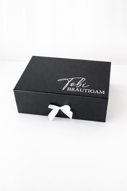 Personalisierte schwarze Geschenkbox für den Bräutigam mit Aufdruck „Tobi Bräutigam“ und weißer Schleife auf hellem neutralen Hintergrund.