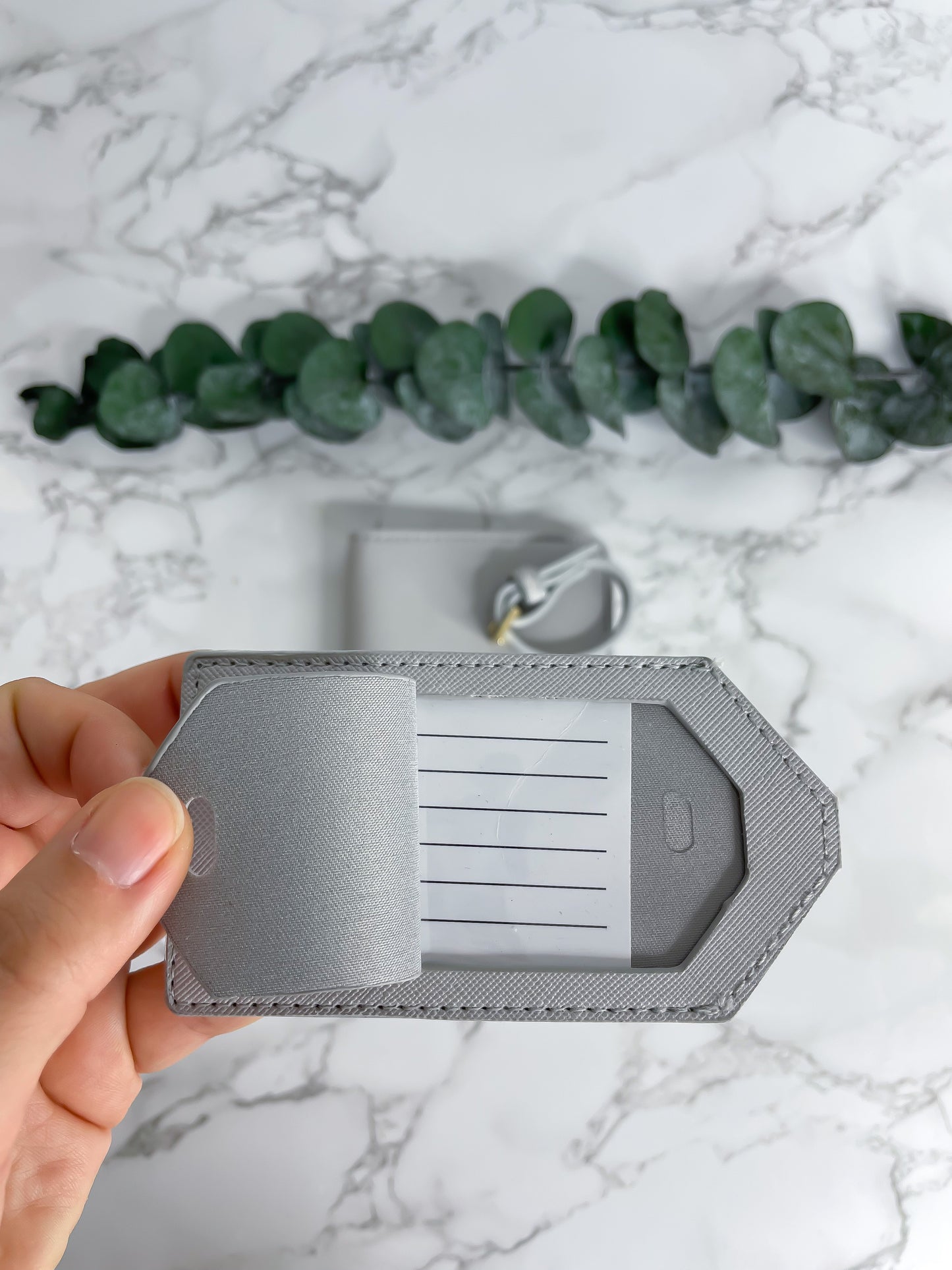 Graue Reisepasshülle aus Saffiano-Leder mit Fächern für Karten und Reisepass sowie passendem Kofferanhänger, dekorativ fotografiert auf Marmor-Hintergrund mit Eukalyptus.