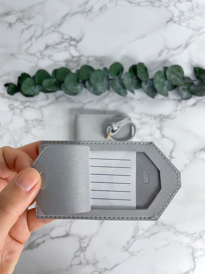 Graue Reisepasshülle aus Saffiano-Leder mit Fächern für Karten und Reisepass sowie passendem Kofferanhänger, dekorativ fotografiert auf Marmor-Hintergrund mit Eukalyptus.