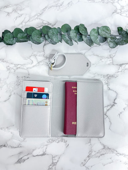 Graue Reisepasshülle aus Saffiano-Leder mit Fächern für Karten und Reisepass sowie passendem Kofferanhänger, dekorativ fotografiert auf Marmor-Hintergrund mit Eukalyptus.