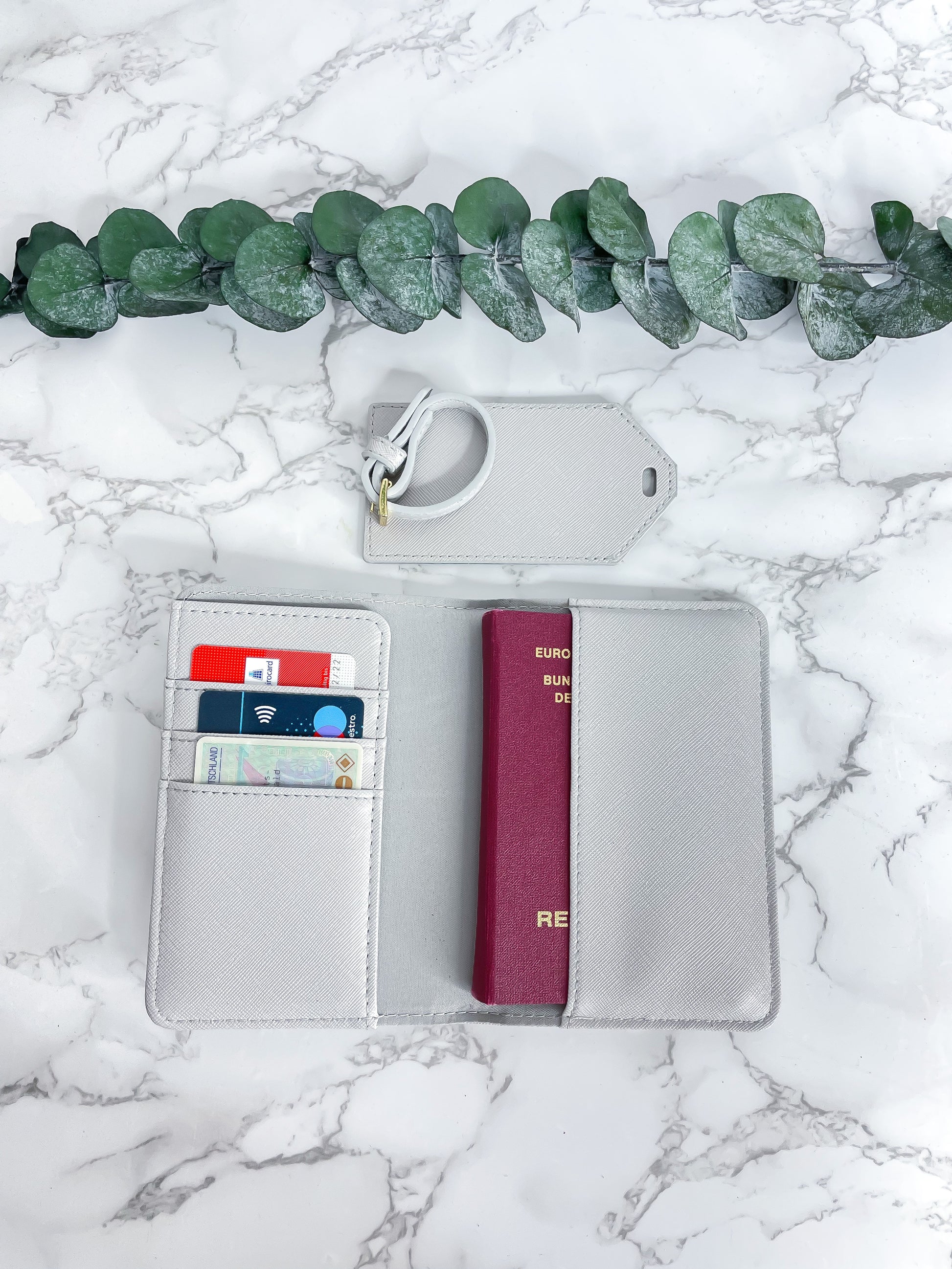 Graue Reisepasshülle aus Saffiano-Leder mit Fächern für Karten und Reisepass sowie passendem Kofferanhänger, dekorativ fotografiert auf Marmor-Hintergrund mit Eukalyptus.